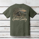An olive green short-sleeve t-shirt shown from the back, featuring a large graphic print of a wild turkey in a scenic landscape with the text 'Gobbler Country' in a distressed beige font. The shirt is displayed flat against a white-washed wooden plank background.
