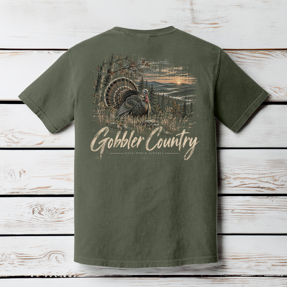 An olive green short-sleeve t-shirt shown from the back, featuring a large graphic print of a wild turkey in a scenic landscape with the text 'Gobbler Country' in a distressed beige font. The shirt is displayed flat against a white-washed wooden plank background.