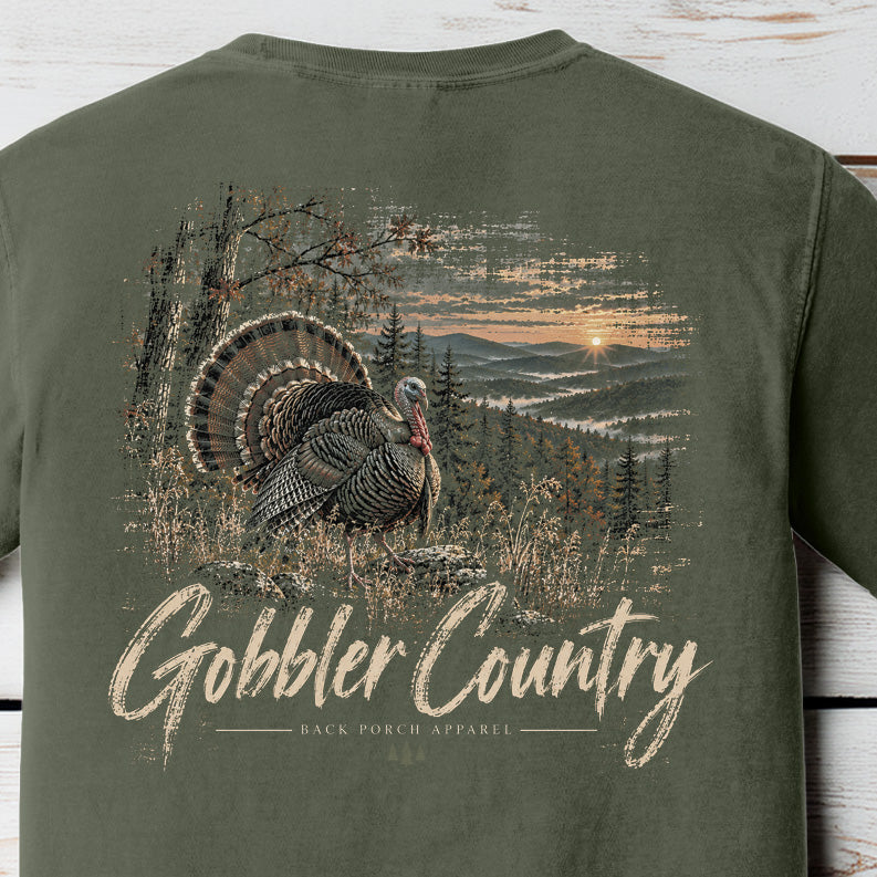 An olive green short-sleeve t-shirt shown from the back, featuring a large graphic print of a wild turkey in a forest landscape with the text 'Gobbler Country' in a distressed beige font.