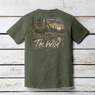 An olive green short-sleeve t-shirt shown from the back, featuring a large graphic print of a moose in a forest by a lake with the text 'The Wild' and 'Black Forest Apparel' printed below it. The shirt is displayed flat against a white-washed wooden plank background.