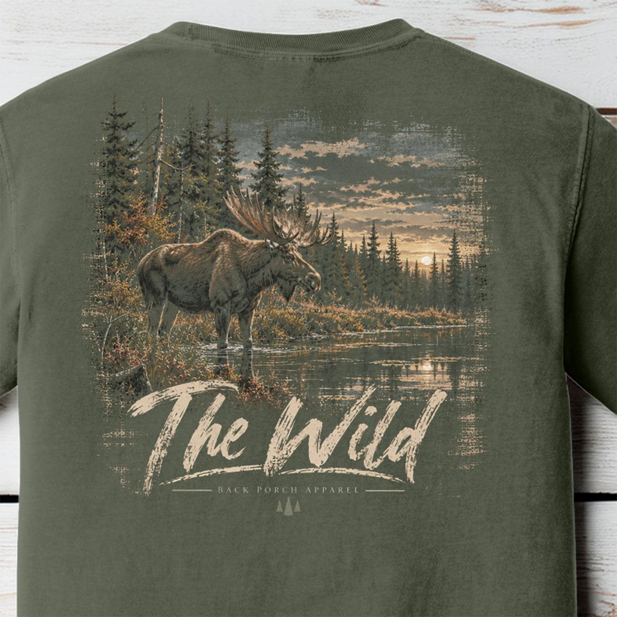 An olive green t-shirt showing the back graphic print with a moose in a forest by a river and the text 'The Wild' and 'Back Forch Apparel'.