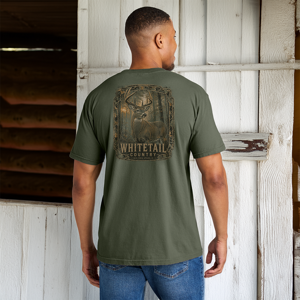 Man wearing the Whitetail Country Comfort Colors Moss t-shirt featuring a trophy whitetail buck standing outside a rustic barn.