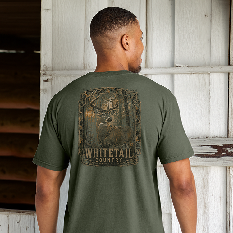 Man wearing the Whitetail Country Comfort Colors Moss t-shirt featuring a trophy whitetail buck standing outside a rustic barn.