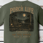 Close-up of the Porch Life Fireflies Are Better on the Porch artwork showing a rocking chair, lantern, moonlit lake, and glowing fireflies on a Moss Comfort Colors t-shirt.