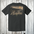 A dark charcoal gray t-shirt hanging on a wooden hanger against a light gray wood plank wall. The back of the shirt features a large graphic print of a sunset over a forested hillside, with the word 'Acadia' in a stylized beige script and a small brown bear logo below.