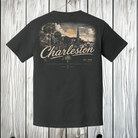 A dark charcoal gray t-shirt shown from the back, featuring a vintage-style graphic of Charleston, SC with a sunset, church steeple, and the word 'Charleston' in cream lettering. The t-shirt is displayed hanging against a white-washed wood plank background.
