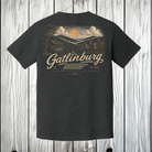 A dark charcoal gray t-shirt shown from the back, featuring a large vintage-style scenic graphic of Gatlinburg, Tennessee, with a mountain landscape, cabin, and a yellow sun. The word 'Gatlinburg' is written in a large beige script across the center.