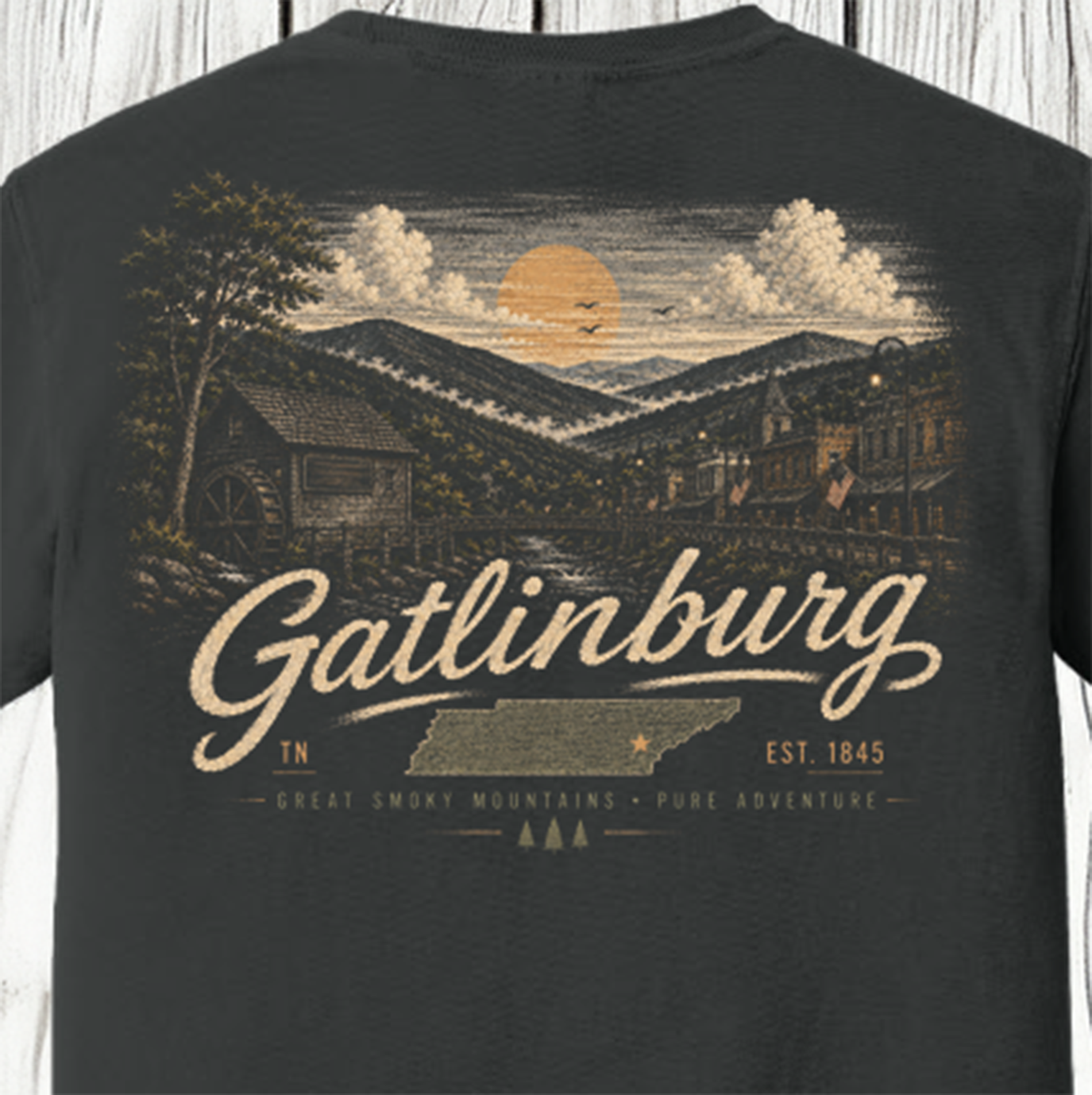 A close-up of the back of a dark charcoal gray t-shirt featuring a detailed vintage-style graphic of Gatlinburg, Tennessee. The graphic depicts a mountain landscape with a waterwheel mill, a town, clouds, and a large yellow sun, with 'Gatlinburg' written in a large cream-colored script font.
