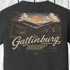 A close-up of the back of a dark charcoal gray t-shirt featuring a detailed vintage-style graphic of Gatlinburg, Tennessee. The graphic depicts a mountain landscape with a waterwheel mill, a town, clouds, and a large yellow sun, with 'Gatlinburg' written in a large cream-colored script font.