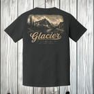 A black short-sleeve t-shirt shown from the back, featuring a large vintage-style graphic of Glacier National Park with mountains, trees, and a lake, printed in beige and brown tones. The t-shirt is hanging on a wooden hanger against a light gray wood plank background.