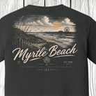 A dark charcoal gray t-shirt featuring a vintage-style graphic print of Myrtle Beach, SC. The design shows a beach sunset scene with a Ferris wheel, dunes, and a palm tree, with 'Myrtle Beach' written in large white script across the front.