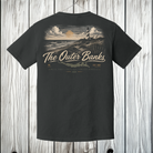 A black short-sleeve t-shirt shown from the back, hanging against a white wood plank background. The shirt features a large graphic print of a lighthouse on a rocky coastline at sunset, with the text 'The Outer Banks' and 'N.