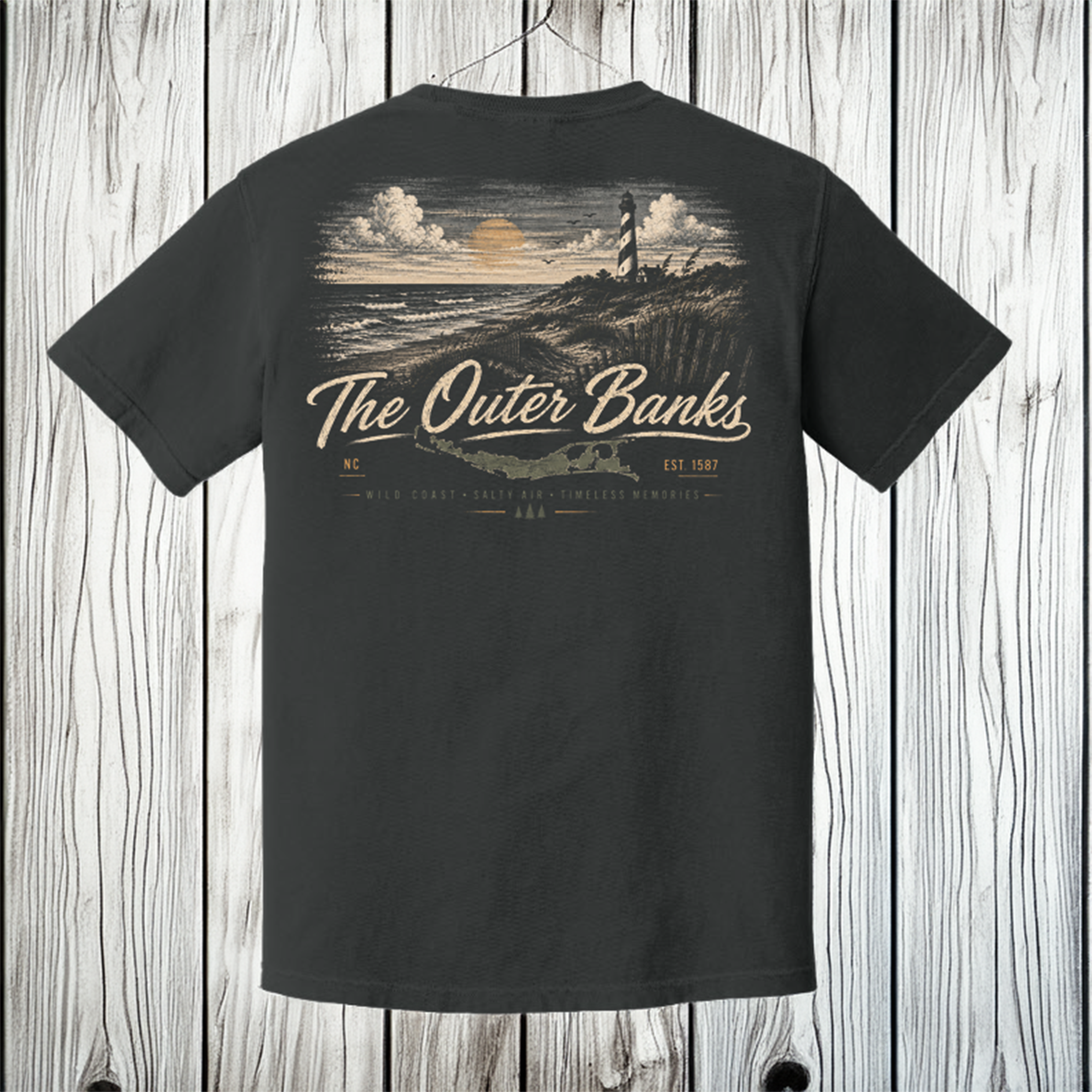 A black short-sleeve t-shirt shown from the back, hanging against a white wood plank background. The shirt features a large graphic print of a lighthouse on a rocky coastline at sunset, with the text 'The Outer Banks' and 'N.