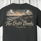 A dark charcoal gray t-shirt featuring a vintage-style graphic print of a lighthouse on a beach under a sunset sky, with the text 'The Outer Banks' and 'NC EST. 1587' printed on the front.
