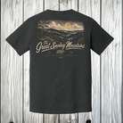 A dark charcoal gray t-shirt hanging on a wooden hanger against a light gray wood plank wall. The back of the shirt features a large graphic print of a sunset over a forested hillside, with the word 'Acadia' in a stylized beige script and a small brown bear logo below.