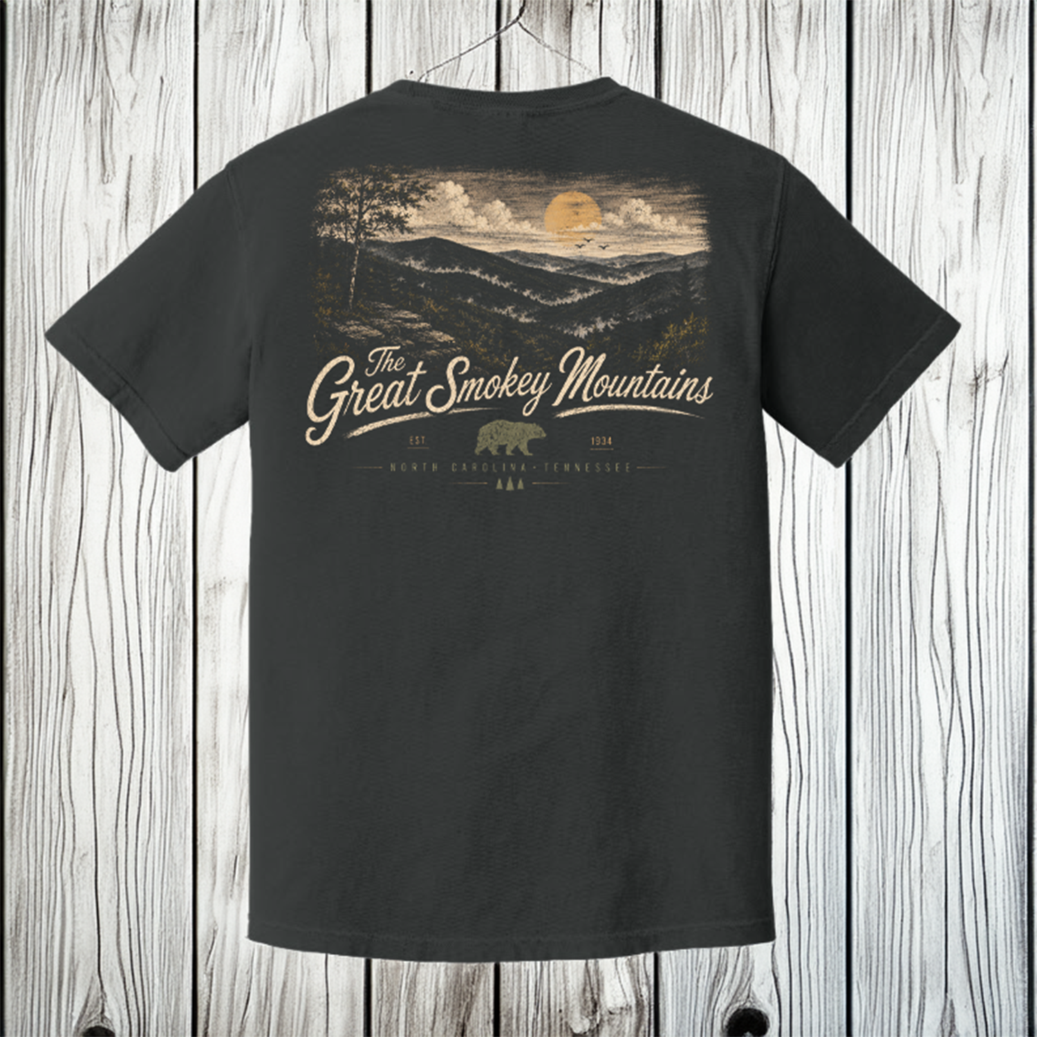 A dark charcoal gray t-shirt hanging on a wooden hanger against a light gray wood plank wall. The back of the shirt features a large graphic print of a sunset over a forested hillside, with the word 'Acadia' in a stylized beige script and a small brown bear logo below.