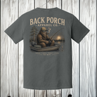 Pepper Comfort Colors Bigfoot fishing t-shirt displayed on a white wood background featuring a peaceful lake fishing scene.