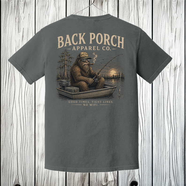 Pepper Comfort Colors Bigfoot fishing t-shirt displayed on a white wood background featuring a peaceful lake fishing scene.