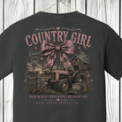 Distressed country girl graphic featuring a pink camouflage bow, vintage tractor, rustic barn, and southern farmhouse artwork on a Comfort Colors Pepper shirt.