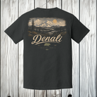 A dark charcoal gray t-shirt with a large back graphic showing a mountain landscape, pine trees, a bear, and the text 'Denali' and 'Alaska 2024', displayed against a rustic white wood plank background.