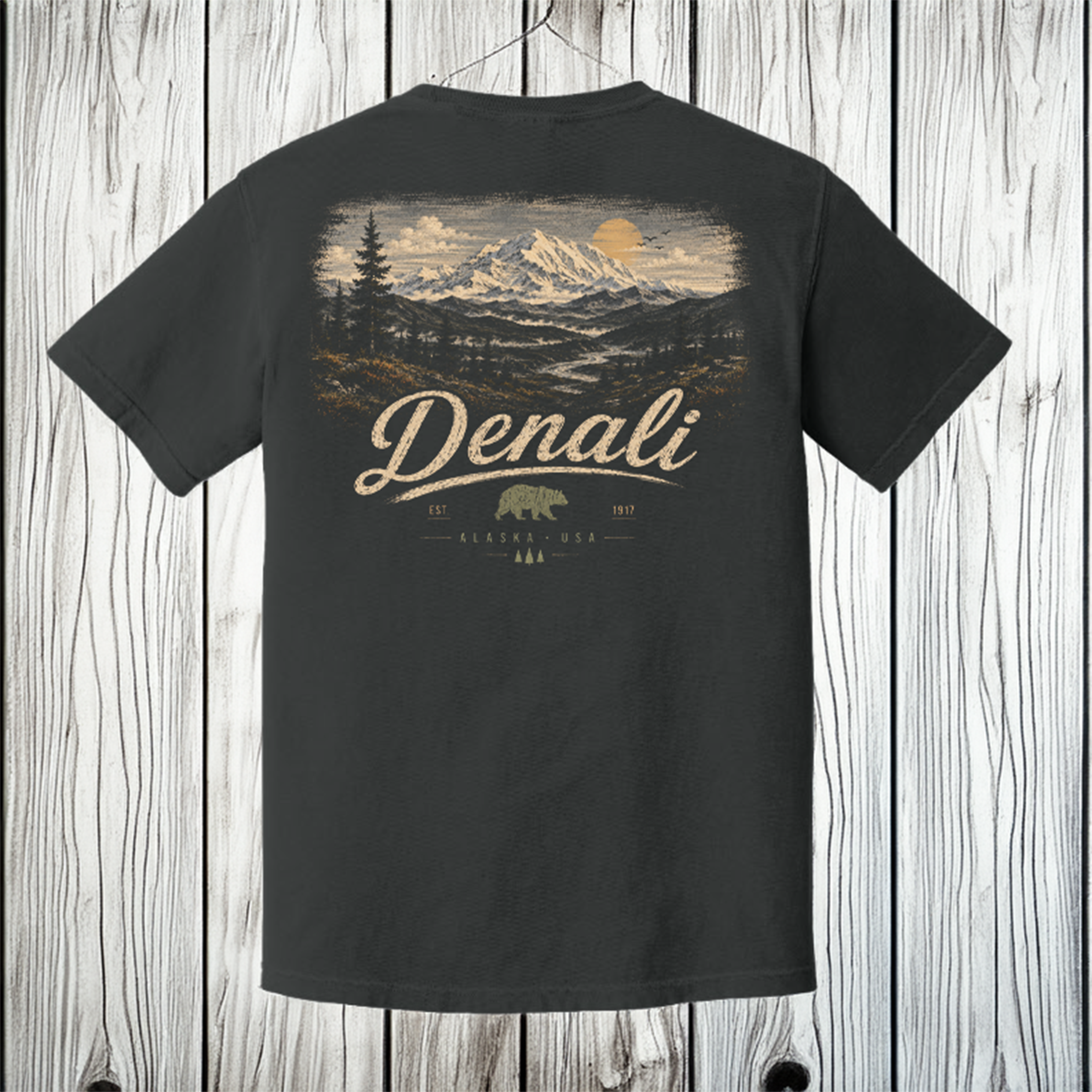 A dark charcoal gray t-shirt with a large back graphic showing a mountain landscape, pine trees, a bear, and the text 'Denali' and 'Alaska 2024', displayed against a rustic white wood plank background.
