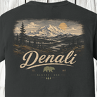 A close-up of a dark charcoal gray t-shirt showing the back side with a detailed graphic print of a mountain landscape and the word 'Denali' in cream-colored lettering.