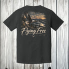 A dark charcoal gray t-shirt with a large graphic print on the back, featuring a bald eagle flying over a lake at sunset with the text 'Flying Free' and 'Mendota Wilderness'. The shirt is displayed against a rustic white-washed wood plank background.