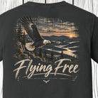 A dark charcoal gray t-shirt showing the back graphic print. The design features a bald eagle flying over a mountain lake at sunset, with the text 'Flying Free' and 'BACK FOR A APPAREL' printed below.