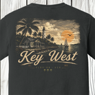 A dark charcoal gray t-shirt featuring a vintage-style graphic print of Key West, Florida. The print depicts a lighthouse, palm trees, a sailboat, and a sunset in muted beige, orange, and brown tones, with 'Key West' written in a large cursive font.