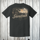 A dark charcoal gray t-shirt shown from the back, featuring a vintage-style graphic of Savannah, Georgia, with a sunset, bridge, and the word 'Savannah' in large script. The shirt is displayed hanging against a white wood plank background.
