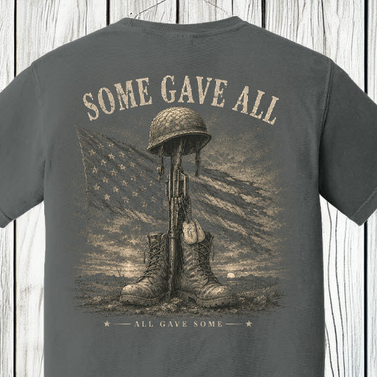 Distressed patriotic military memorial graphic featuring combat boots, rifle, helmet, dog tags, and an American flag on a Comfort Colors Pepper shirt.