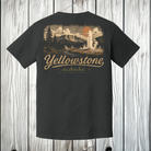 A dark charcoal gray t-shirt shown from the back, featuring a large vintage-style Yellowstone National Park graphic in beige and orange. The shirt is hanging on a wooden hanger against a light gray wood plank background.