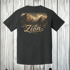 A dark charcoal gray t-shirt shown from the back, featuring a vintage-style graphic of Zion National Park with the word 'Zion' in a large cursive font. The t-shirt is displayed hanging against a rustic white-washed wooden plank background.