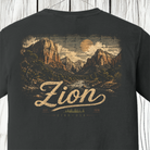 A dark charcoal gray t-shirt featuring a vintage-style scenic graphic of Zion National Park with a large textured 'Zion' script and 'UT - USA' text below, displayed on a wooden plank background.