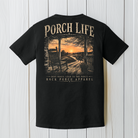 Black Comfort Colors Porch Life graphic t-shirt displayed against a rustic wood background featuring a peaceful country porch at sunset.
