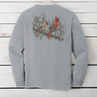 A light gray long-sleeve t-shirt shown from the back, featuring a graphic print of two red cardinals perched on a snowy branch. The shirt is laid flat on a white wooden plank background.