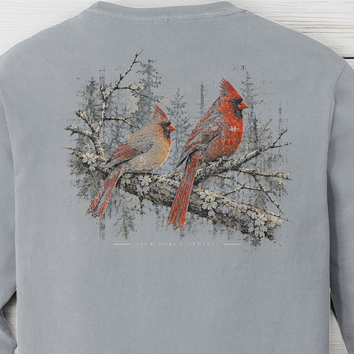 A light gray long-sleeve crewneck sweatshirt featuring a large graphic print of two red cardinal birds perched on a flowering branch with a snowy forest background.