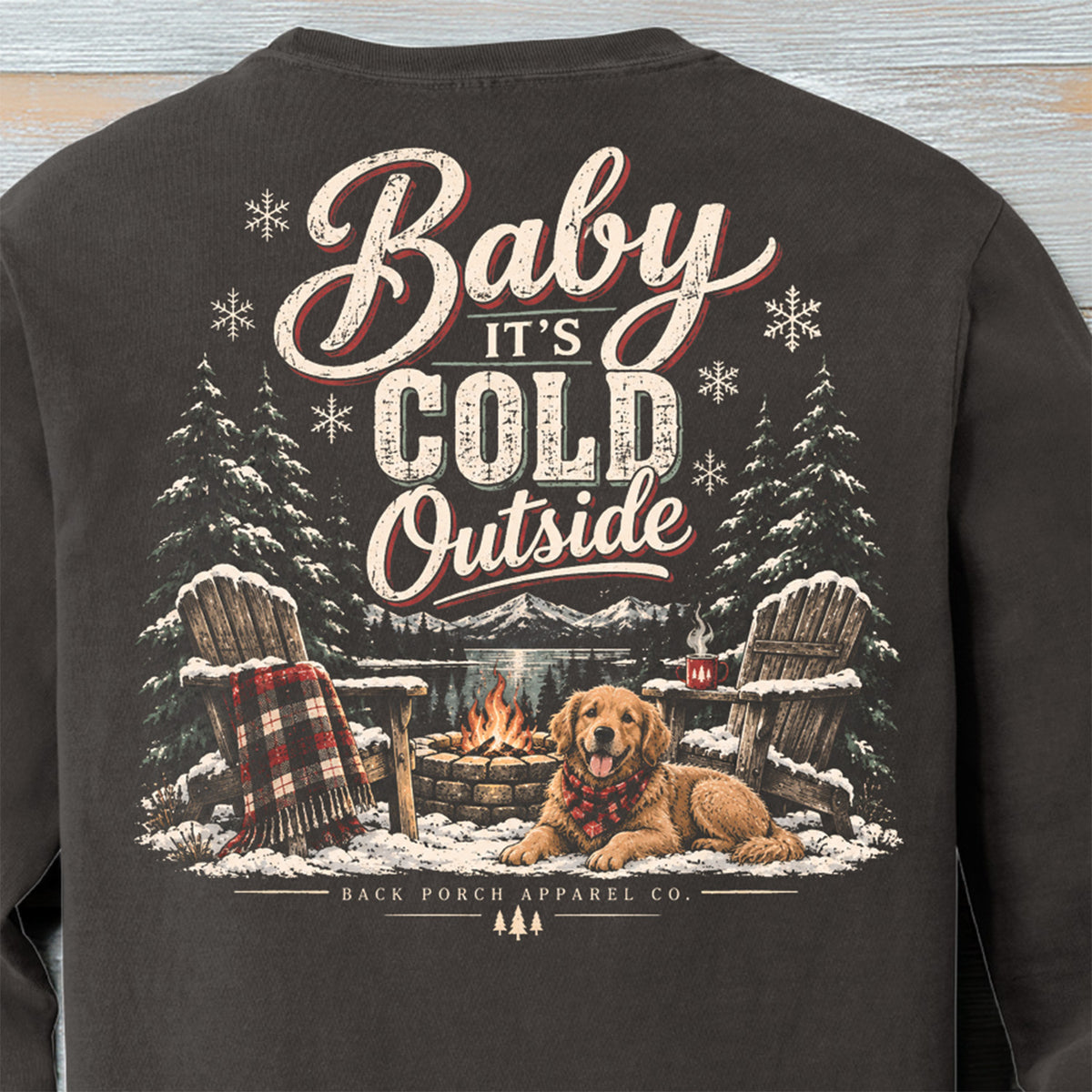 Close-up of Baby It’s Cold Outside winter shirt design with golden retriever, campfire, Adirondack chairs, snowy pines and mountains