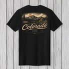 A black short-sleeve t-shirt with a vintage-style Colorado mountain landscape graphic on the back, featuring the text 'Colorado' in a beige script font, displayed against a light gray wood plank background.