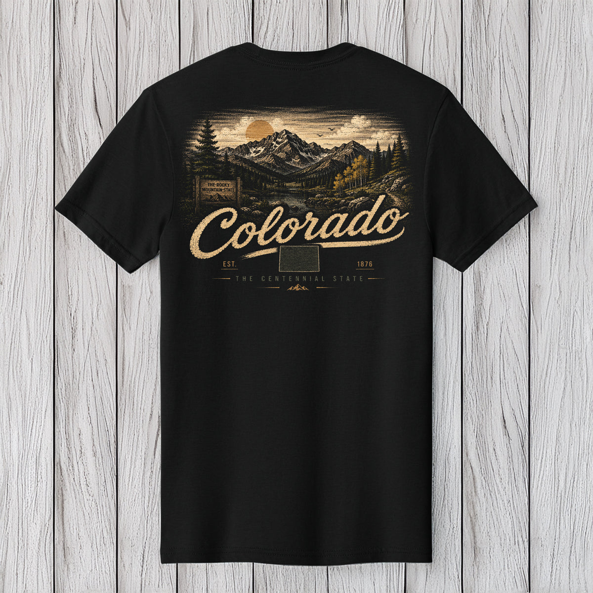 A black short-sleeve t-shirt with a vintage-style Colorado mountain landscape graphic on the back, featuring the text 'Colorado' in a beige script font, displayed against a light gray wood plank background.