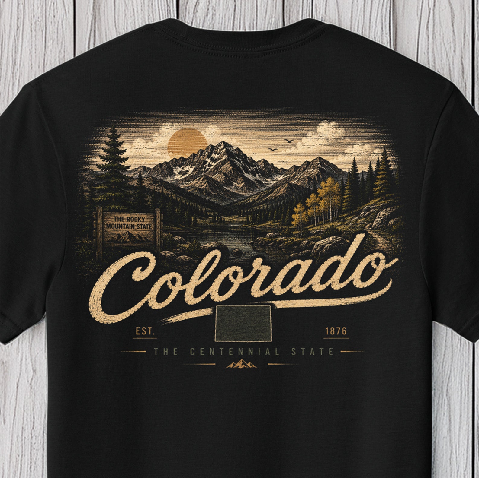 A black short-sleeve t-shirt featuring a vintage-style graphic print of a Colorado mountain landscape with a river, trees, and a large 'Colorado' script across the front, displayed against a light gray wood plank background.
