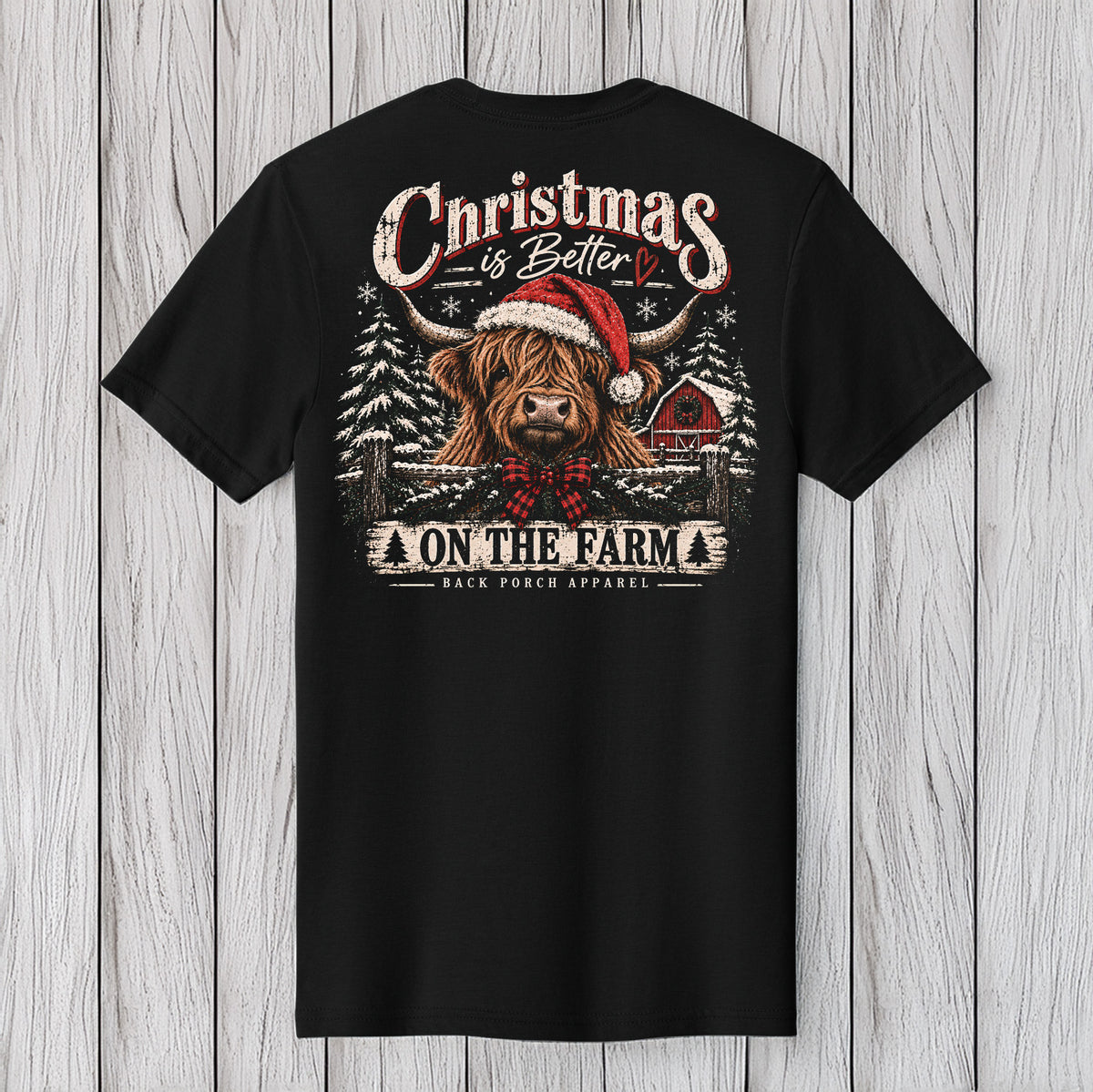 A black short-sleeve t-shirt displayed flat against a light gray wood plank background, showing the back graphic. The graphic features a Highland cow in a red Santa hat and plaid bow tie, with the text 'Christmas is Better ON THE FARM' and 'BLACK DOGS APPAREL' printed below.