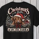 A black short-sleeve graphic t-shirt displayed flat against a light gray wood plank background. The shirt features a festive Christmas design with a highland cow wearing a red Santa hat and red plaid bow tie, surrounded by snow-covered pine trees, a red barn, and distressed text reading 'Christmas is Better ON THE FARM' with 'BACK PORCH APPAREL' printed below.
