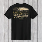 A black short-sleeve t-shirt with a vintage-style Kentucky landscape graphic on the front, featuring a barn, trees, and a winding road under a sunset sky, with 'Kentucky' written in large cream script. The t-shirt is displayed flat against a light gray wood plank background.