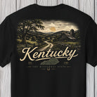 A black short-sleeve t-shirt with a vintage-style graphic on the back depicting a Kentucky countryside scene with a dirt road, barn, silo, and trees. The words 'Kentucky', 'EST.