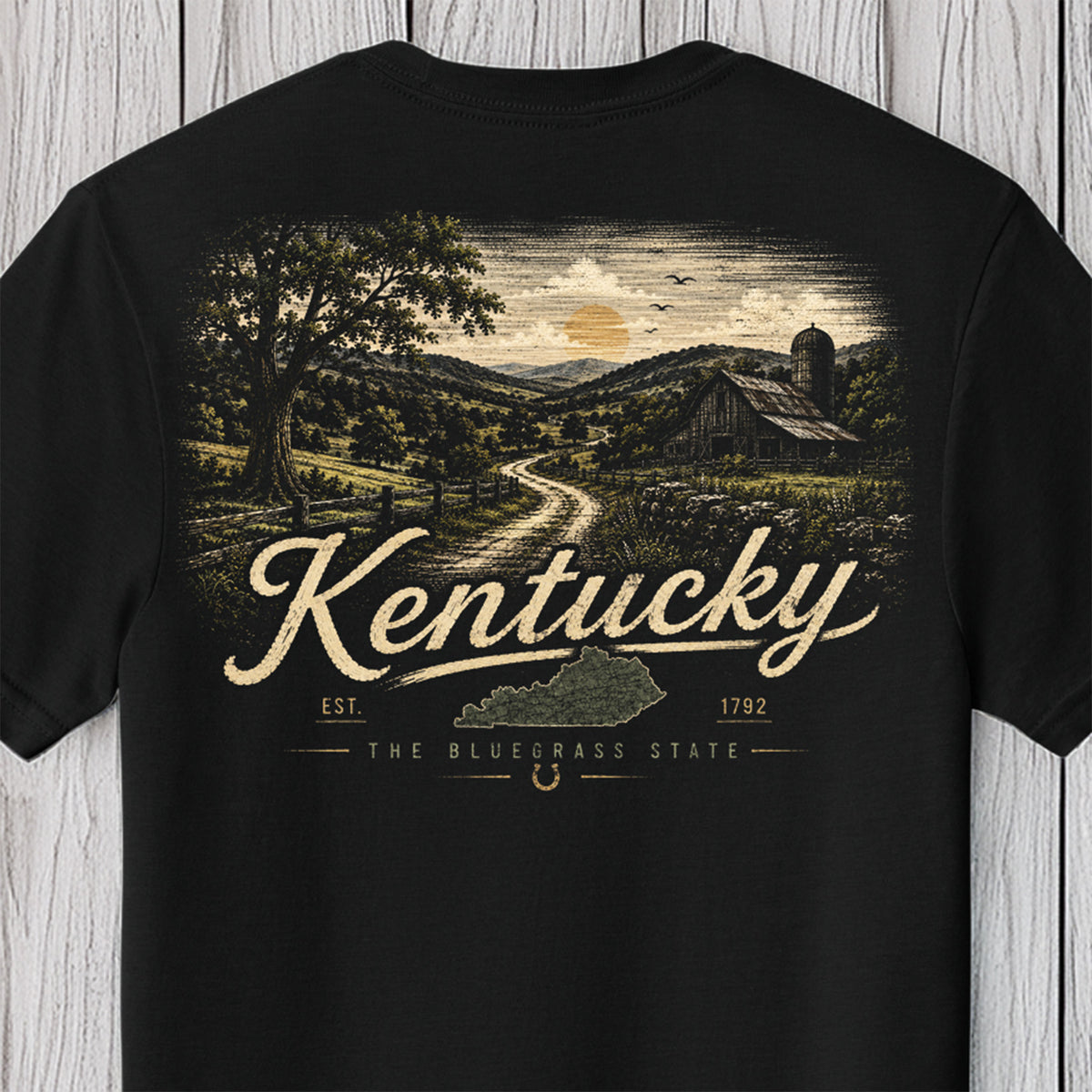 A black short-sleeve t-shirt with a vintage-style graphic on the back depicting a Kentucky countryside scene with a dirt road, barn, silo, and trees. The words 'Kentucky', 'EST.