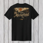 A black short-sleeve t-shirt with a vintage-style Maryland graphic on the back, displayed against a light gray wood plank background.