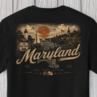 A black short-sleeve t-shirt with a detailed vintage-style graphic of Maryland, featuring a lighthouse, sailboats on the water, a large orange sun, and the word 'Maryland' in script across the front.
