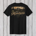 A black short-sleeve t-shirt with a vintage-style Montana landscape graphic on the back, featuring mountains, trees, and the word 'Montana' in a tan script, displayed against a light gray wood plank background.