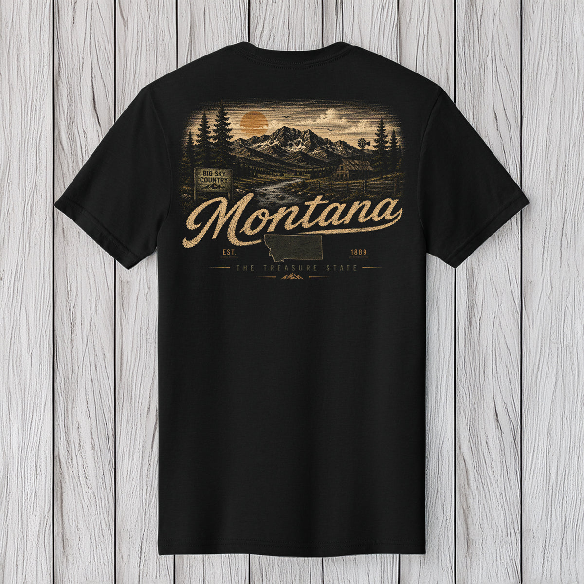 A black short-sleeve t-shirt with a vintage-style Montana landscape graphic on the back, featuring mountains, trees, and the word 'Montana' in a tan script, displayed against a light gray wood plank background.